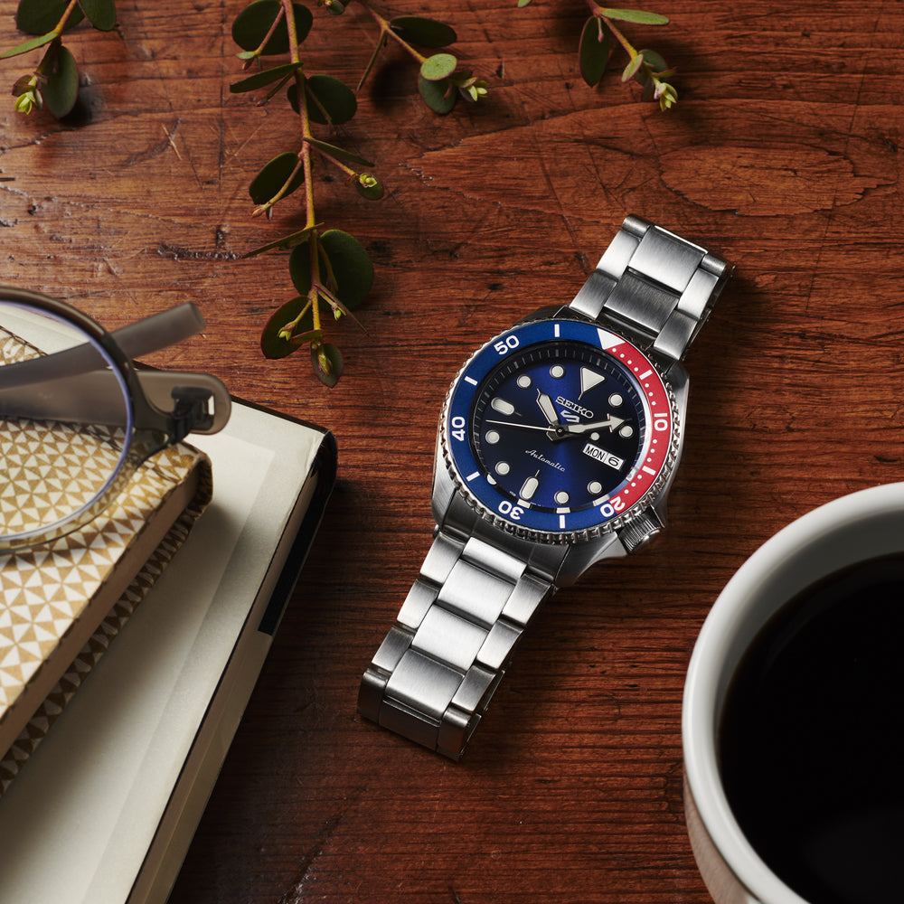 Een Seiko 5 Sports heren horloge SRPD53K1 van Seiko, met een blauwe en rode rand, ligt op een houten tafel naast een koffiekopje, een bril, een notitieboekje met patroon en een takje met bladeren.