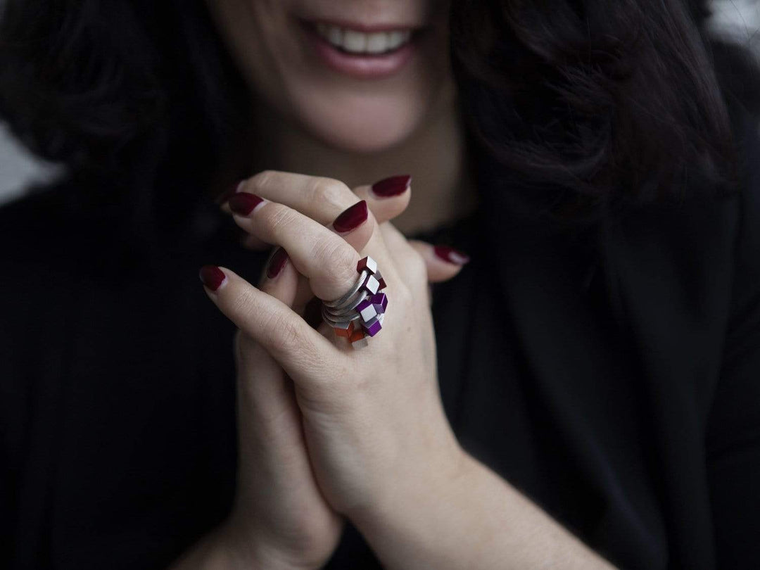 Een vrouw met donker haar lacht, haar handen gevouwen onthullen donkerrode nagellak en een grote CLIC Creations Ring Kubusjes Oranje R4O met een uniek ontwerp met paarse en roze stenen. Ze is gekleed in het zwart.