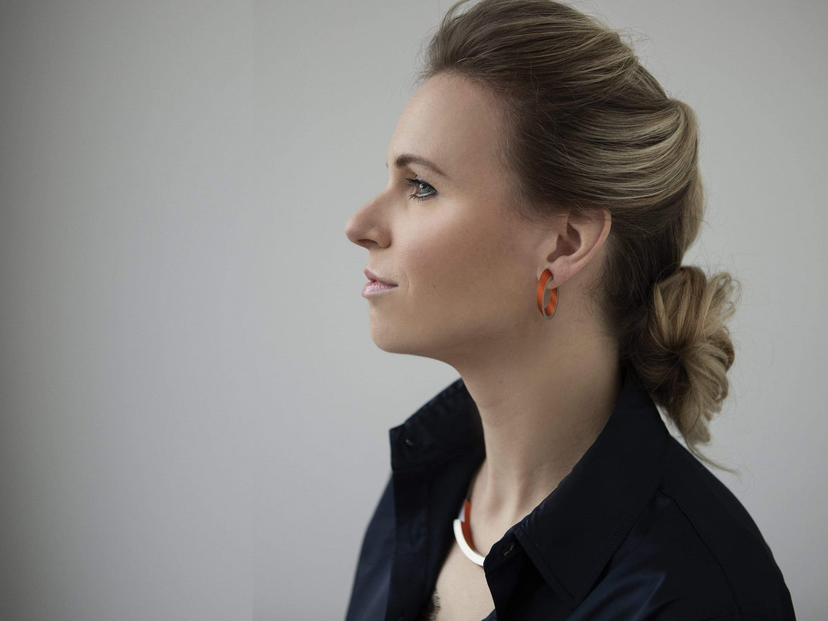 Een vrouw met licht haar in een lage knot en oranje oorbellen draagt een donker overhemd met kraag, aangevuld met de Collier Bogen Rood C74R van CLIC Creations. Ze kijkt naar links tegen een neutrale achtergrond.