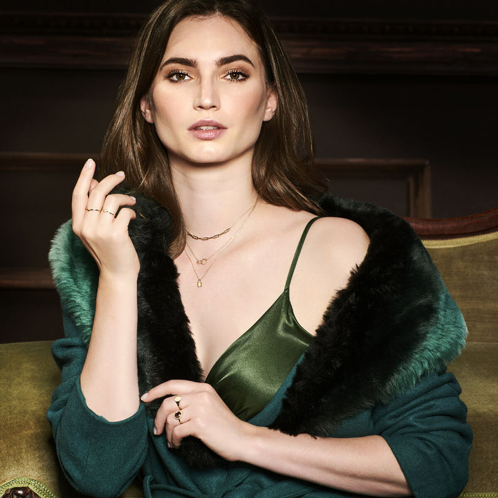 Een vrouw met lang bruin haar draagt een groene satijnen jurk en een donkergroene jas met bontrand, zittend op een groene stoel. Ze draagt gouden ringen, waaronder de Van der Kooij Collectie ring 14K geelgoud, en kijkt zelfverzekerd in de camera.