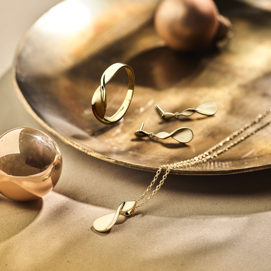 Een Van der Kooij Collectie ring poli/mat 14K geelgoud, oorstekers en een hangerketting met een eenvoudige, gedraaide elegantie worden gepresenteerd op een metalen schaal met decoratieve bollen, badend in warm, zacht licht.