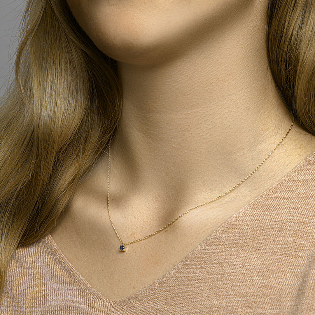 Een vrouw draagt een beige top met V-hals en een Van der Kooij Collectie Gouden dames ketting saffier 14K, met een kleine ronde hanger. Alleen haar nek, ondergezicht en een deel van haar haar zijn zichtbaar.