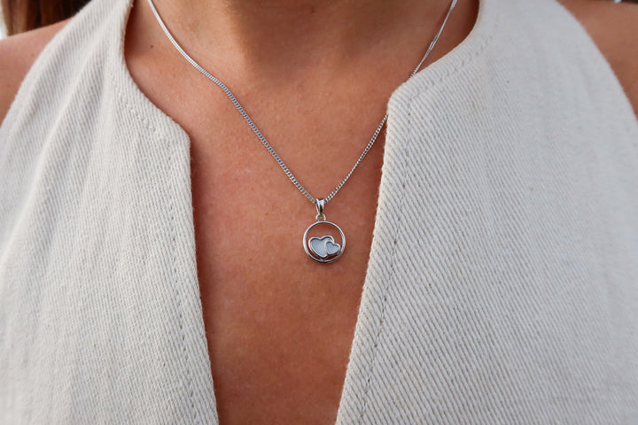 Een persoon draagt een mouwloze top met crèmekleurige textuur en de Amélie AsHanger massief halsketting van Juwelier van der Kooij, met een ronde hanger met hartmotief. De focus ligt op de ketting en de bovenkant van de borst.