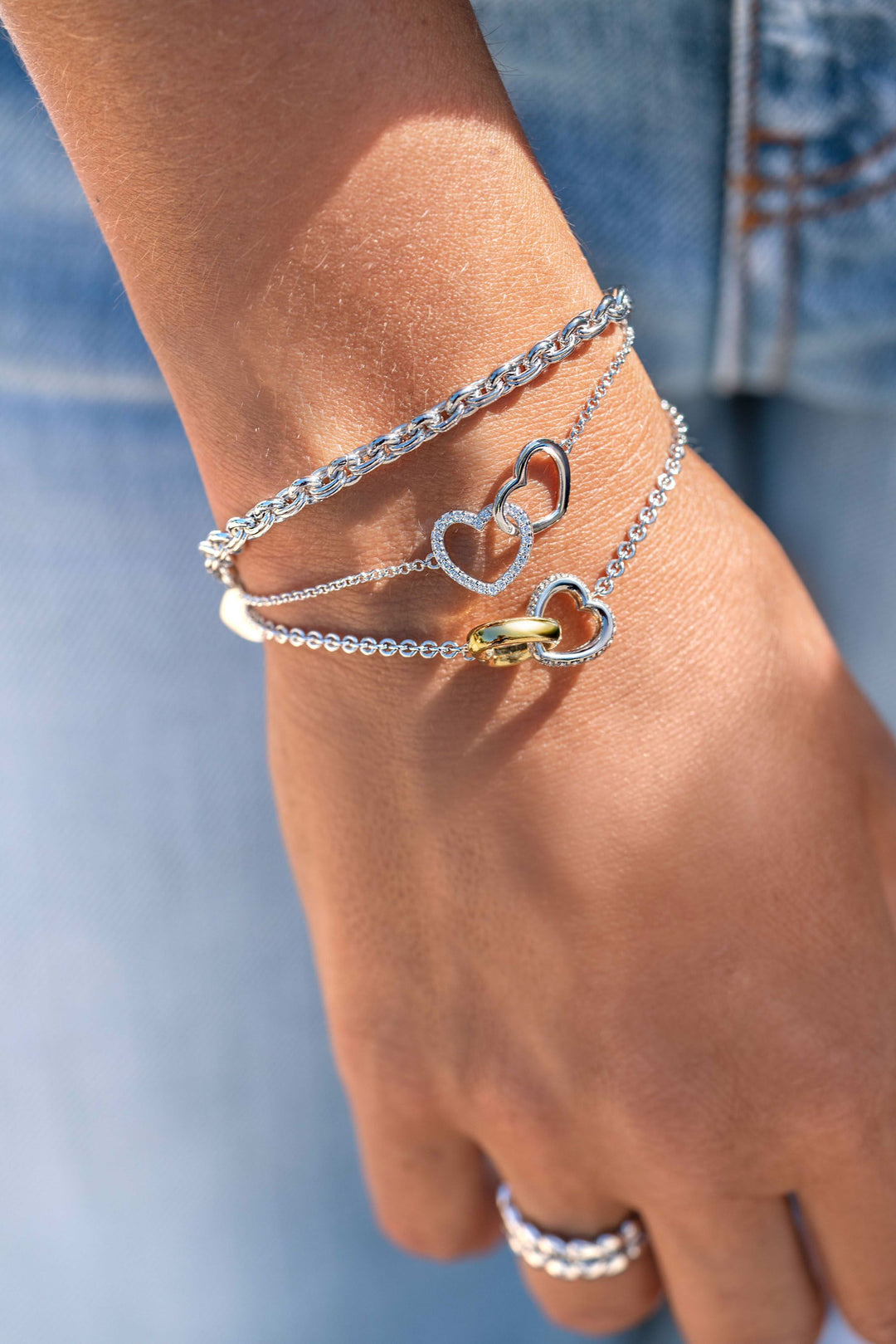 Een hand versierd met drie zilveren Zinzi-armbanden met hartontwerpen en een Zinzi Armband Zilver Zia1911, gecombineerd met een glimmende zilveren ring, past perfect bij een spijkerbroek, omdat de sieraden schitterend glinsteren in het licht.