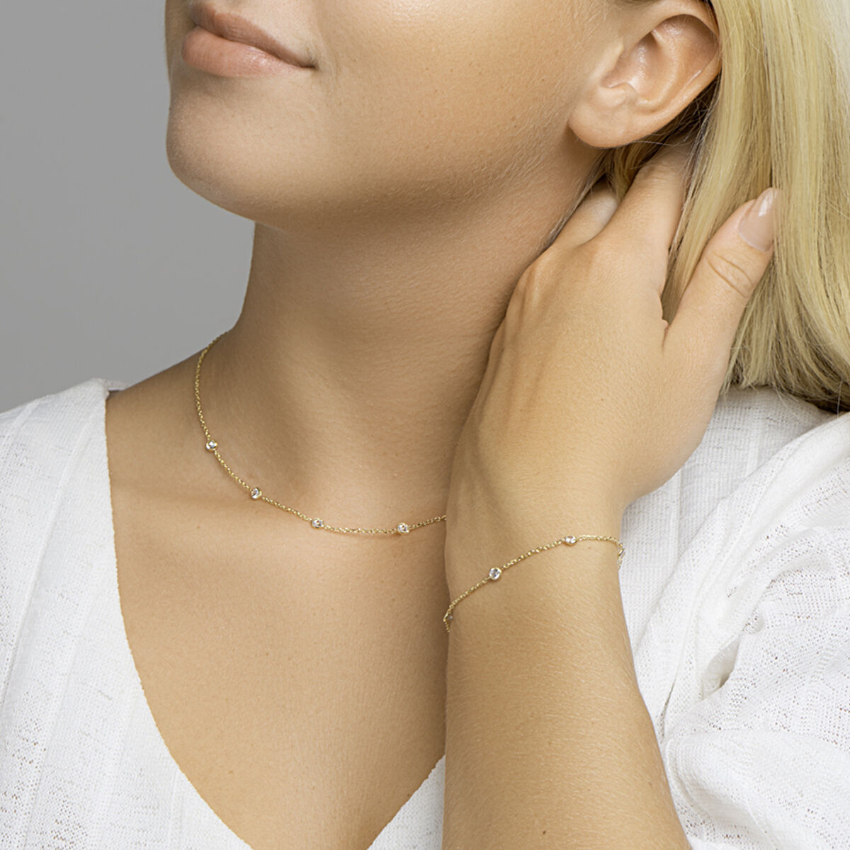 Een vrouw in een witte top met V-hals strijkt zachtjes over haar hals, draagt een gouden halsketting en de Van der Kooij Collectie armband zirkonia 17 + 2 cm 3 micron zilver verguld (geel)- het perfecte accessoire voor stijlvolle dames.