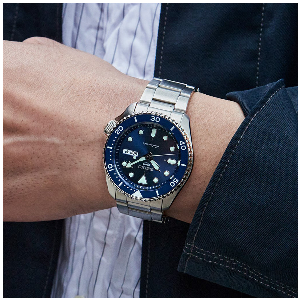 Een persoon verbetert zijn look met een Seiko 5 Sports heren horloge SRPD51K1 met een zilveren metalen band en blauwe wijzerplaat, die de tijd, secondemarkeringen en de datum weergeeft. Ze combineren het met een donker jasje over een gestreept shirt.