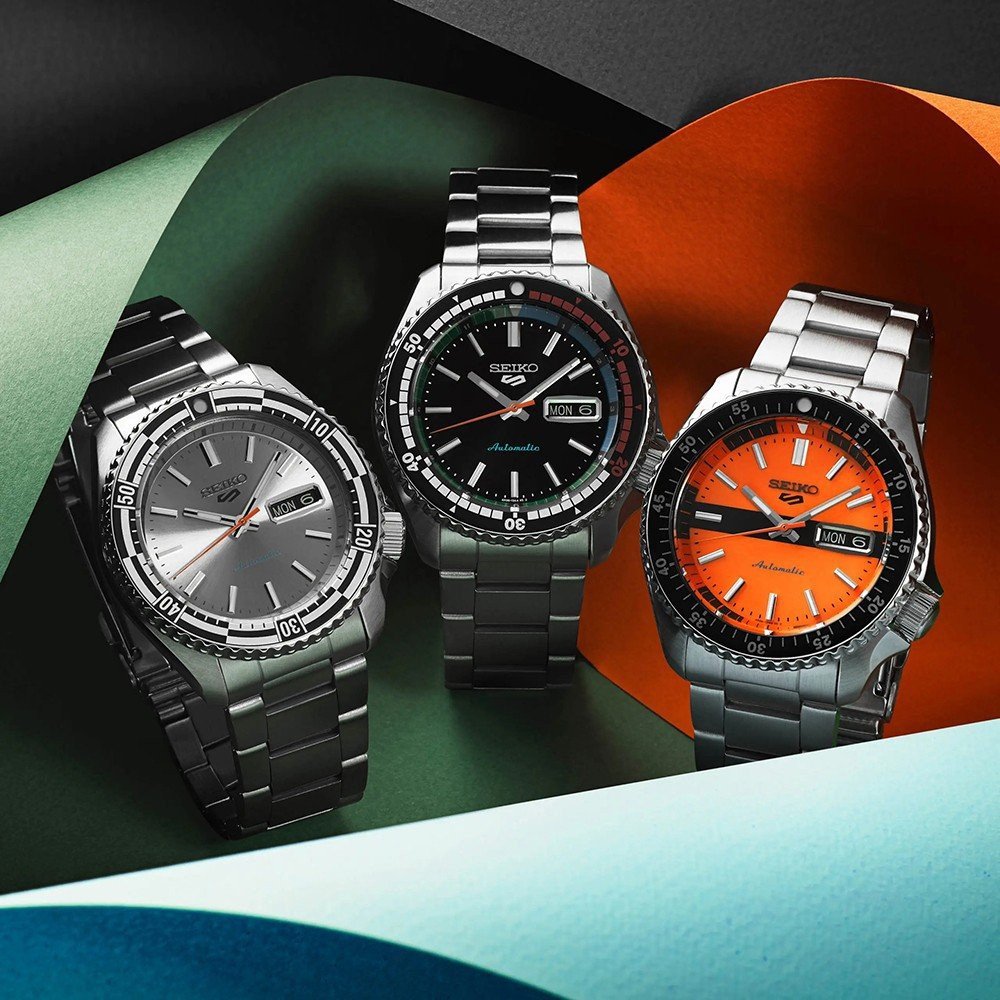 Drie Seiko 5 Sports SRPK11K1 horloges met metalen banden staan tegen een kleurrijke achtergrond. De zilveren, zwarte en oranje wijzerplaten hebben allemaal datum- en dagvensters. Met een waterdichtheid van 10ATM benadrukt het display het ontwerp en de duurzaamheid van de automatische uurwerken.