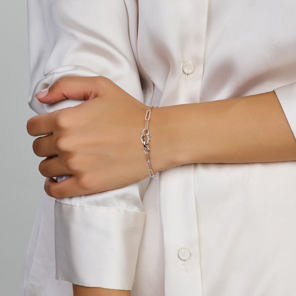 Iemand draagt een wit satijnen overhemd met lange mouwen en draagt de Van der Kooij Collectie armband paperclip platte buis 3,8 mm 17 cm met groot springslot van gerhodineerd zilver om zijn pols.