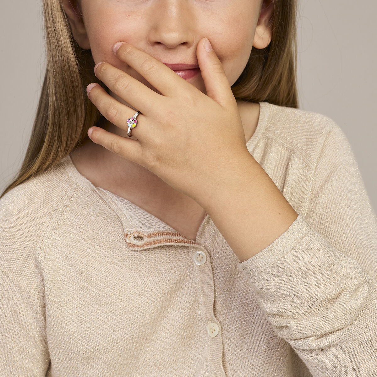 Een jong meisje met lang haar glimlacht vriendelijk en houdt haar hand voor haar mond. Ze draagt een beige vest en een Van der Kooij Collectie ring met bloem en zirkonia zilver gerhodineerd aan haar vinger. De achtergrond is neutraal.