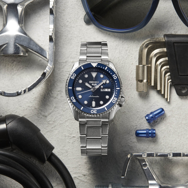 De Seiko 5 Sports heren horloge SRPD51K1, een zilveren horloge met een blauwe wijzerplaat en metalen band, wordt weergegeven op een gestructureerd oppervlak, vergezeld door een zonnebril, een moersleutel, inbussleutels, een kabel en twee blauwe doppen. Dit automatische horloge toont elegant zowel de tijd als de datum.