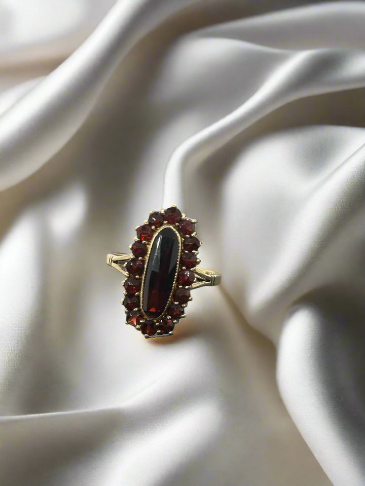Een vintage gouden ring 14k ovale granaat 3,7 gram van Juwelier van der Kooij, met een langwerpige donkerrode edelsteen omringd door kleinere rode stenen, ligt op zacht gevouwen witte satijnen stof, onderdeel van hun Vintage Collectie, en creëert een elegante en luxueuze achtergrond.