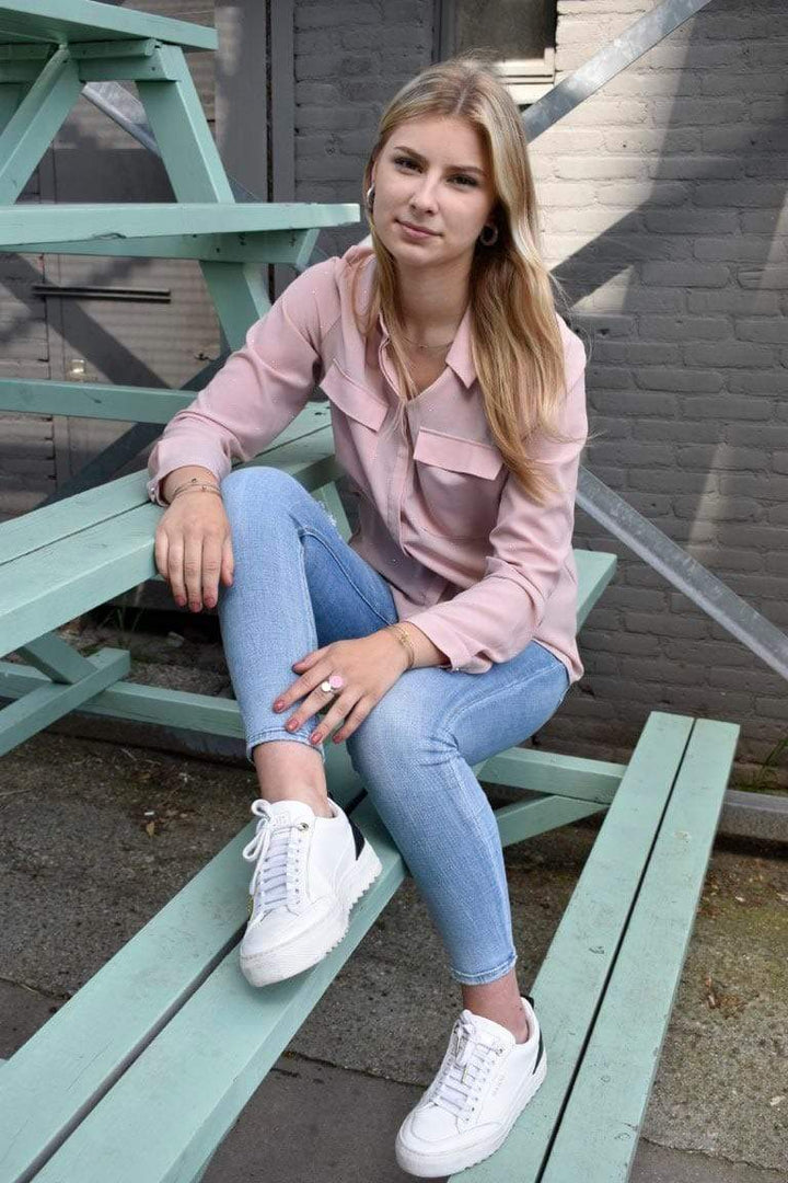 Een persoon met lang, blond haar zit nonchalant op mintgroene houten treden, draagt CLIC Creations Ring Twee ronde vormen Roze R5RO en een lichtroze overhemd. Hun neutrale uitdrukking voegt charme toe aan de grijze bakstenen achtergrond, aangevuld met lichtblauwe jeans en witte sneakers.