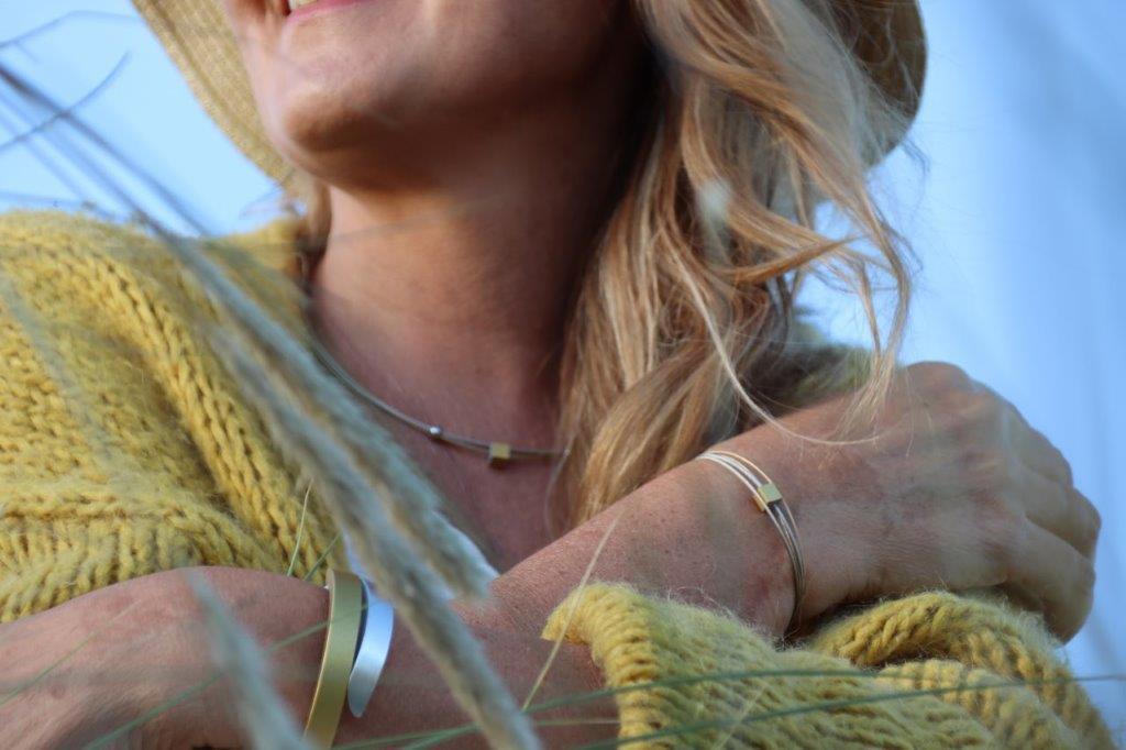 Een vrouw in een gele gebreide trui en muts lacht zachtjes, haar blonde haar valt naar één kant. Ze draagt verschillende sieraden, waaronder de Collier Klein Kubusje Oranje C230O van CLIC Creations. Grassprietjes verschijnen op de voorgrond tegen een zachtblauwe achtergrond.