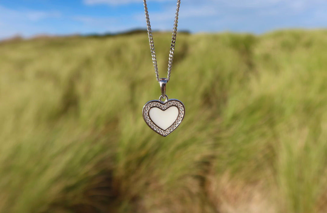 De Demi | AsHanger massief | Zirkonia van Juwelier van der Kooij heeft een zilveren hanger in de vorm van een hart met zirkonia's en een wit hart. De hanger staat scherp af tegen het hoge groene gras en een blauwe lucht.
