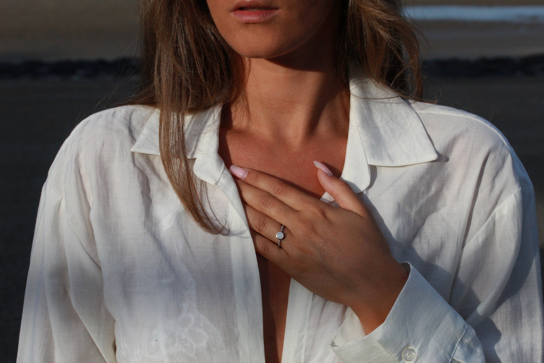 Een vrouw in een gedeeltelijk open witte blouse raakt haar borst aan en toont de See You Isabelle asring zilver met een edelsteen. Haar verzorgde nagels accentueren de elegantie tegen een wazige achtergrond buitenshuis, en belichamen de essentie van de persoonlijke assieraad.