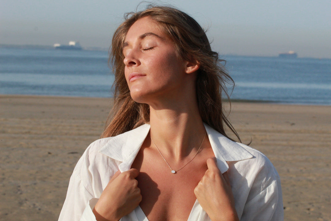 Op een zonnig strand staat een vrouw met haar ogen dicht, de oceaan achter haar. Een wit shirt valt over haar schouders, als aanvulling op haar Luna AsKetting Zilver van See You.