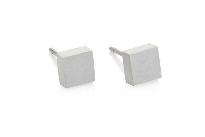 De CLIC Creations Oorsteker Klein vierkant O37 zijn minimalistische vierkante oorstekers, handgemaakt en hypo-allergeen, met een matte metalen afwerking. Deze eenvoudige, geometrische oorknopjes rusten elegant op een reflecterend wit oppervlak.