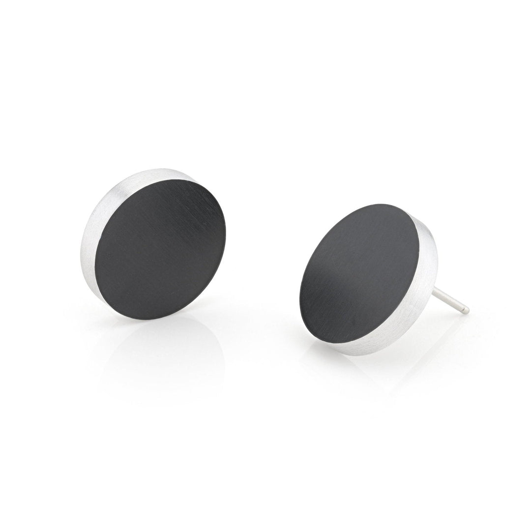De Oorsteker rond O53Z van CLIC Creations, ronde zwarte oorstekers met zilveren randjes, worden op een witte achtergrond getoond. Handgemaakt in Nederland, één oorbel staat rechtop terwijl de andere sierlijk op zijn kant ligt. Het zijn hypo-allergeen oorsieraden.