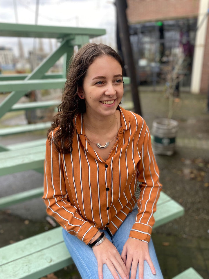 Een persoon met lang bruin haar, gekleed in een oranje gestreept shirt en een blauwe spijkerbroek, zit op mintgroene treden buiten. Ze glimlachen terwijl ze naar de zijkant kijken in een uniek ontworpen omgeving met wazige planten en een gebouw erachter. Ze dragen de Oorsieraad Kubusjes Blauw O28B van CLIC Creations.