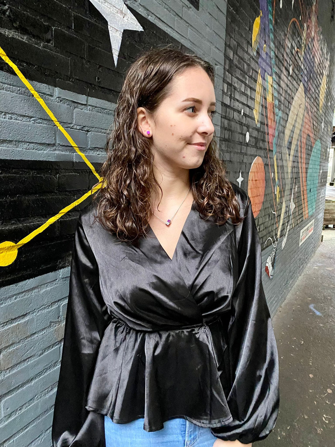 Een vrouw met krullend bruin haar staat tegen een kleurrijke muurschildering, gekleed in een zijdezachte zwarte blouse en blauwe jeans. Ze kijkt opzij en toont haar profiel terwijl ze Collier Twee kubusjes Blauw C205B van CLIC Creations draagt, die perfect past bij het unieke ontwerp van de muurschildering.