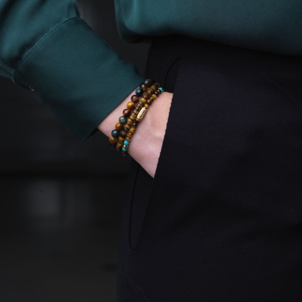 Een persoon in een donkergroen shirt met lange mouwen en een zwarte broek heeft zijn hand in zijn zak en laat verschillende bruine en turquoise kralenarmbanden zien, waaronder de Mix Tiger Turquoise 4mm dames kralen armband van Rebel & Rose om zijn pols.