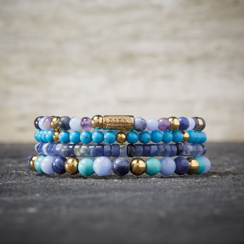 Four Rebel & Rose Zijn viooltjes blauw? 18 kt geelgouden geïoniseerde 4 mm kralenarmbanden voor vrouwen tonen blauwe, paarse, turquoise en gouden kralen die op elkaar gestapeld zijn op een donker gestructureerd oppervlak met een neutrale achtergrond.