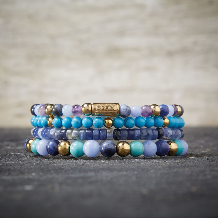 Een stapel kralenarmbanden van Rebel & Rose, met de Winter Blues - 6mm - Kralen Armband Dames, wordt weergegeven op een grijs gestructureerd oppervlak tegen een vage beige achtergrond in blauwe, turquoise, paarse en gouden tinten.