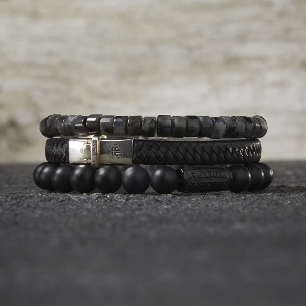Drie luxe sieraden gestapeld: de Rebel & Rose Slices - Grey Seduction - 6mm heren kralen armband, een zwart leren herenarmband met zilveren sluiting en een matte zwarte kralenarmband op een donkere ondergrond.