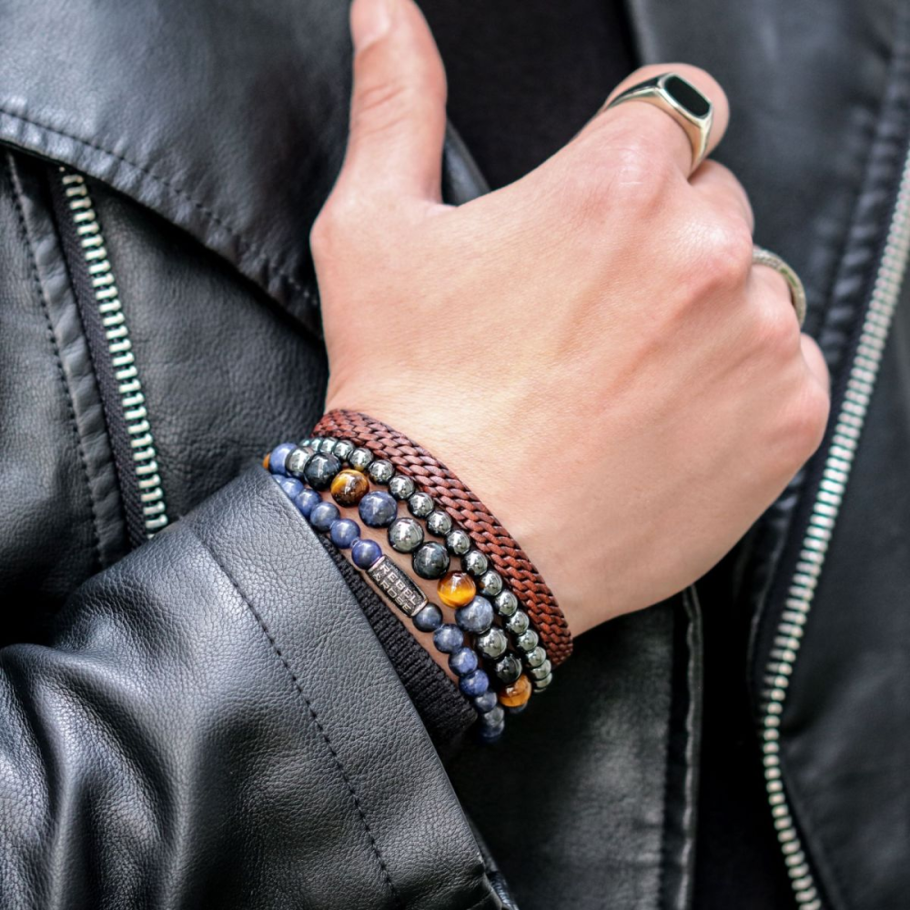 Een persoon in een zwart leren jack toont zijn hand met verschillende kralenarmbanden, waaronder de Rebel & Rose Grey Silver Shine DV - 6mm Kralen Armband Heren, plus een bruin gevlochten band en een dikke zilveren ring.