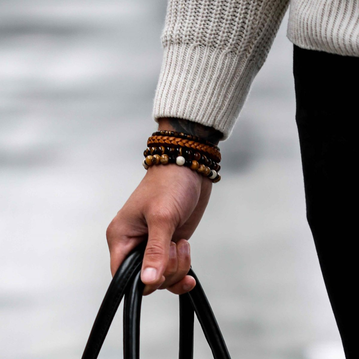 Een persoon in een crèmekleurige gebreide trui en verschillende kralenarmbanden, waaronder de Rebel & Rose kralen Woodstock - 8mm - Kralen Heren Armband, houdt een zwarte tas vast. De achtergrond is onscherp.