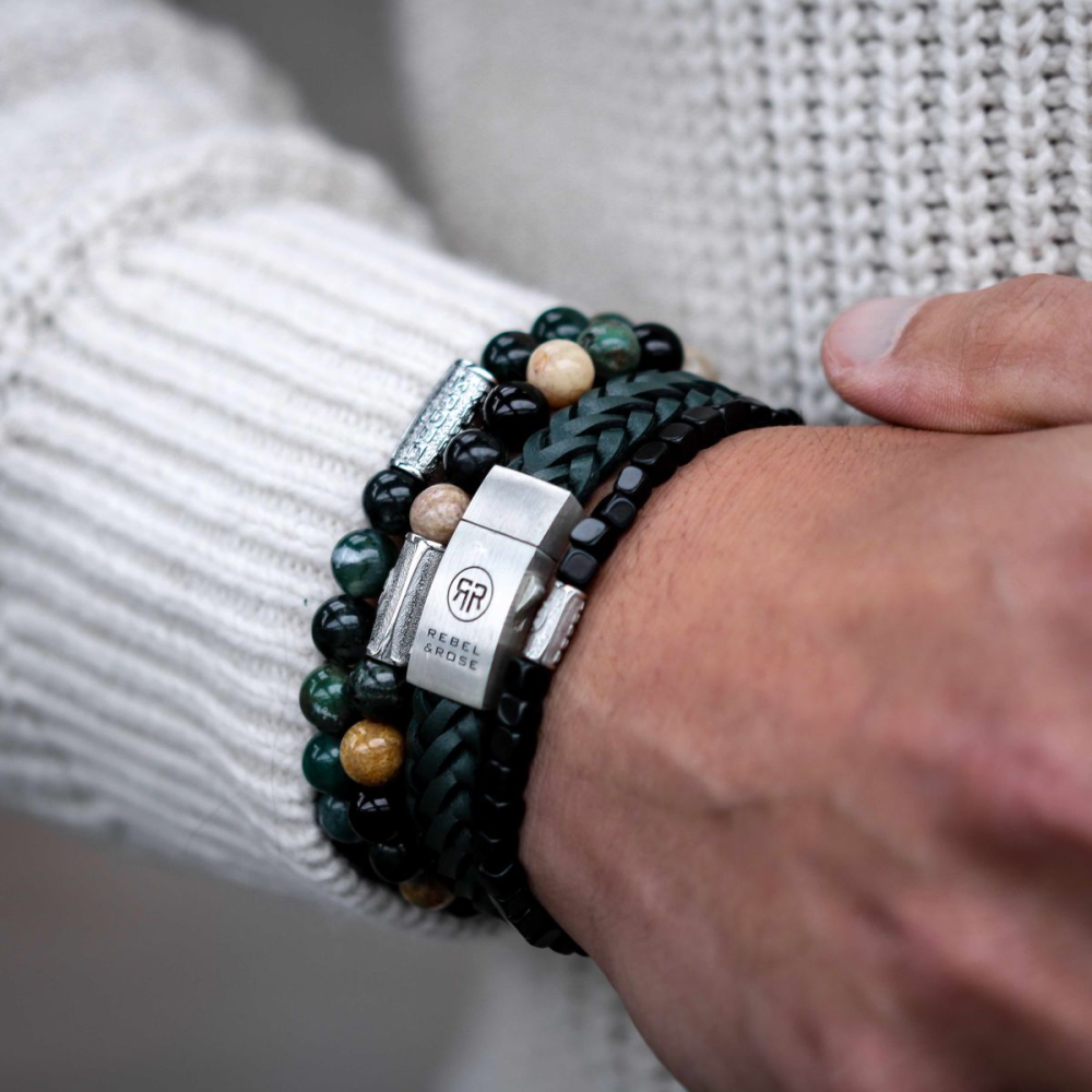 Een persoon in een crèmekleurige gebreide trui omklemt zijn handen en draagt verschillende kralen en gevlochten armbanden, waaronder de Rebel & Rose Braided Raw Matt Ranger leren herenarmband met zilveren sluiting.