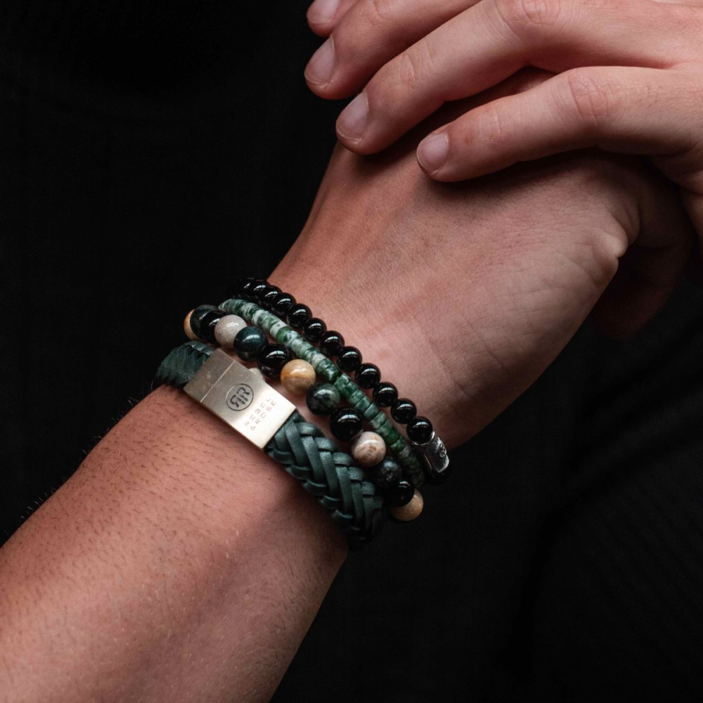 Een close-up van in elkaar geklemde handen accentueert vier armbanden: een Rebel & Rose gevlochten Raw Matt Ranger leren armband met zilveren sluiting, twee kralenarmbanden in zwart en aardetinten en een armband met groene stenen - allemaal om de pols gedragen.