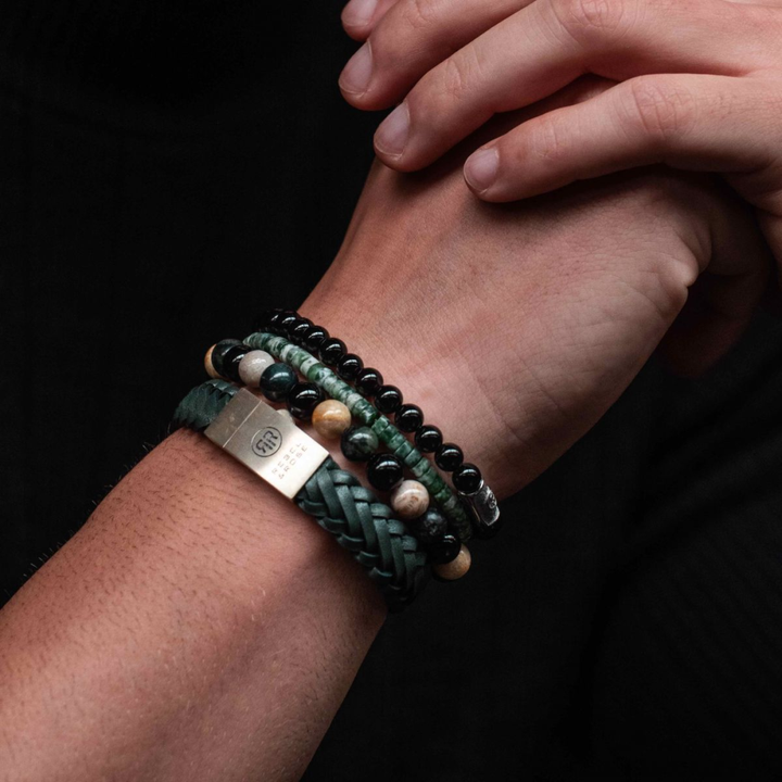 Een close-up van in elkaar geklemde handen accentueert vier armbanden: een Rebel & Rose gevlochten Raw Matt Ranger leren armband met zilveren sluiting, twee kralenarmbanden in zwart en aardetinten en een armband met groene stenen - allemaal om de pols gedragen.
