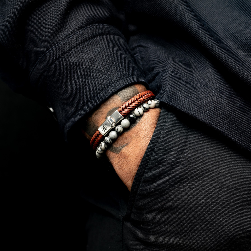 Een hand in een donkere zak, met een zwart overhemd en twee armbanden: een luxe bruin gevlochten leren herenarmband met metalen sluiting, en de Rebel & Rose Black Wolf - 6 mm heren kralen armband met gemarmerde zwarte & grijze agaat kralen.