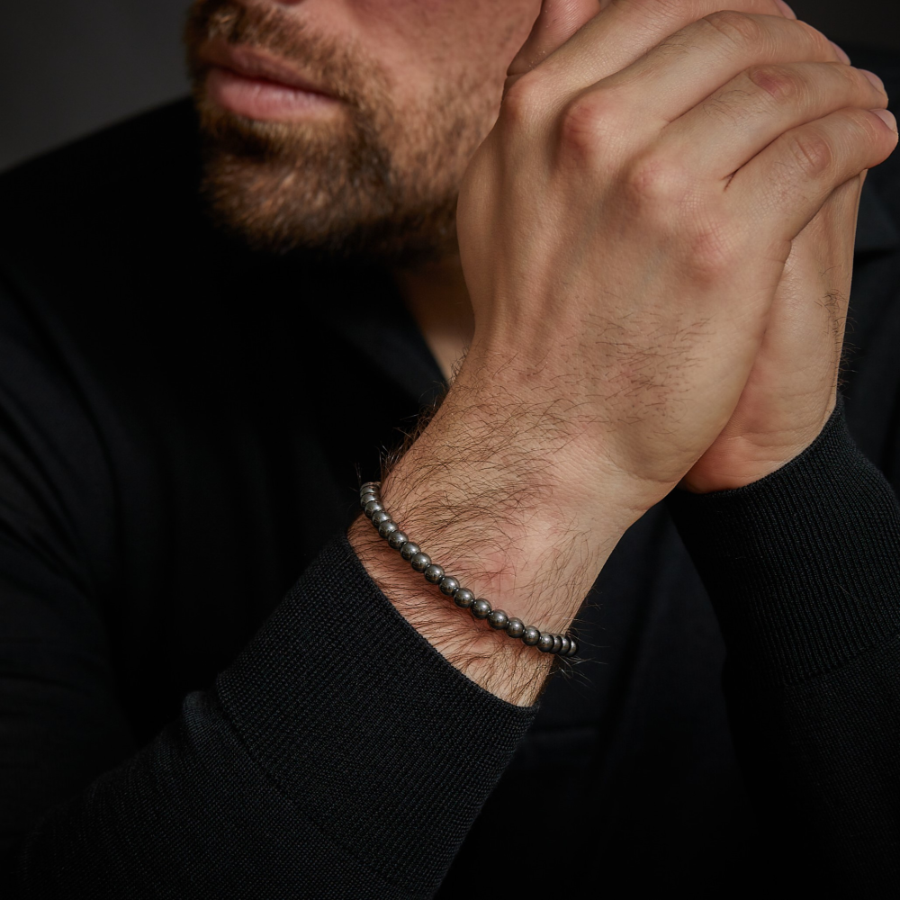 Een man met een getrimde baard draagt een zwart shirt met lange mouwen en omklemt zijn handen terwijl hij de Rebel & Rose Grey Silver Shine DV - 6mm kralen armband heren showt. De afbeelding is bijgesneden om te focussen op zijn ondergezicht, handen en de stijlvolle hematiet armband.