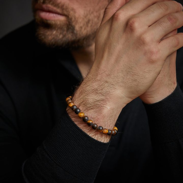 Een bebaarde man in een zwart shirt met lange mouwen omklemt zijn handen en toont de Rebel & Rose Tiger Lily - 8mm Kralen Armband Heren, een stijlvolle herenarmband met bruine en zwarte kralen om zijn pols.