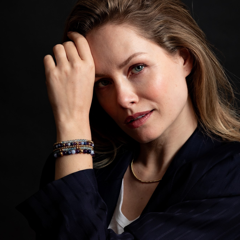 Een vrouw met lichtbruin haar en blauwe ogen laat haar hand op haar voorhoofd rusten. Ze draagt de Rebel & Rose "Are Violets Blue?" 18 kt geelgouden geïoniseerde 4mm armband, een gouden halsketting, een wit topje en een donkere blazer tegen een zwarte achtergrond. 18 kt geelgouden geïoniseerde 4mm kralenarmband, een gouden halsketting, wit topje en donkere blazer tegen een zwarte achtergrond.