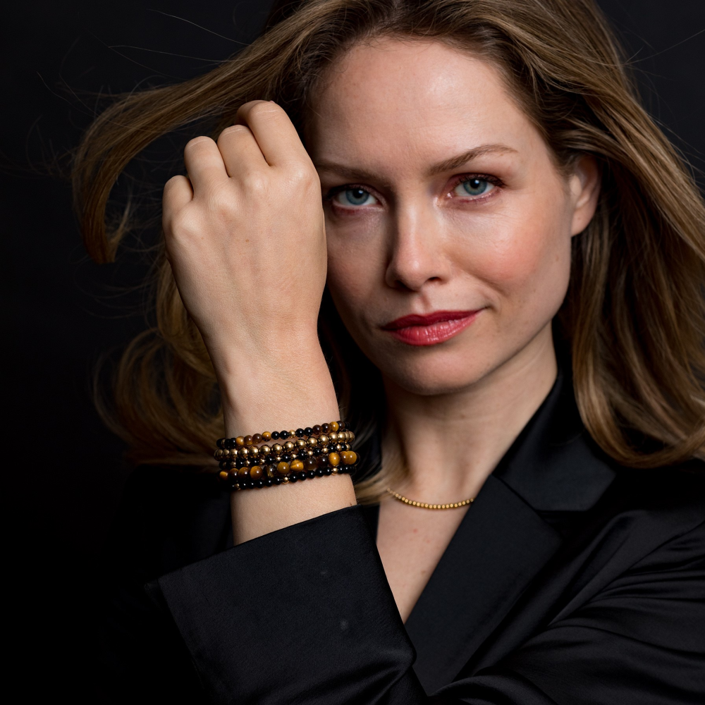 Een vrouw met golvend blond haar, blauwe ogen en een lichte huid kijkt zelfverzekerd naar de camera en toont haar pols versierd met de Rebel & Rose Yellow Gold Only - 6mm Kralen Dames Armband tegen een donkere achtergrond.
