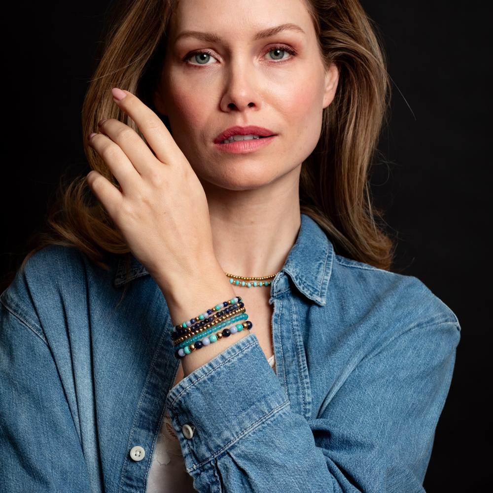 Een vrouw met een denim shirt en kleurrijke kralenarmbanden, waaronder de Rebel & Rose Winter Blues 18 kt geelgouden geïoniseerde 4mm kralen armband dames, poseert zelfverzekerd tegen een donkere achtergrond met één hand bij haar gezicht.