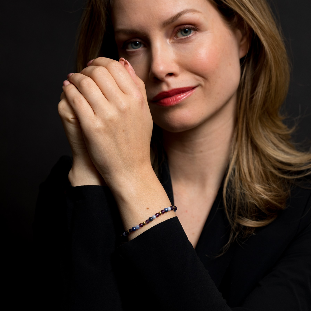 Een vrouw met lichtbruin haar en een lichte huid draagt een zwart topje en de Rebel & Rose "Are Violets Blue?" 18 kt geelgouden geïoniseerde 4mm kralenarmband, poserend met haar handen dicht bij haar gezicht tegen een donkere achtergrond.