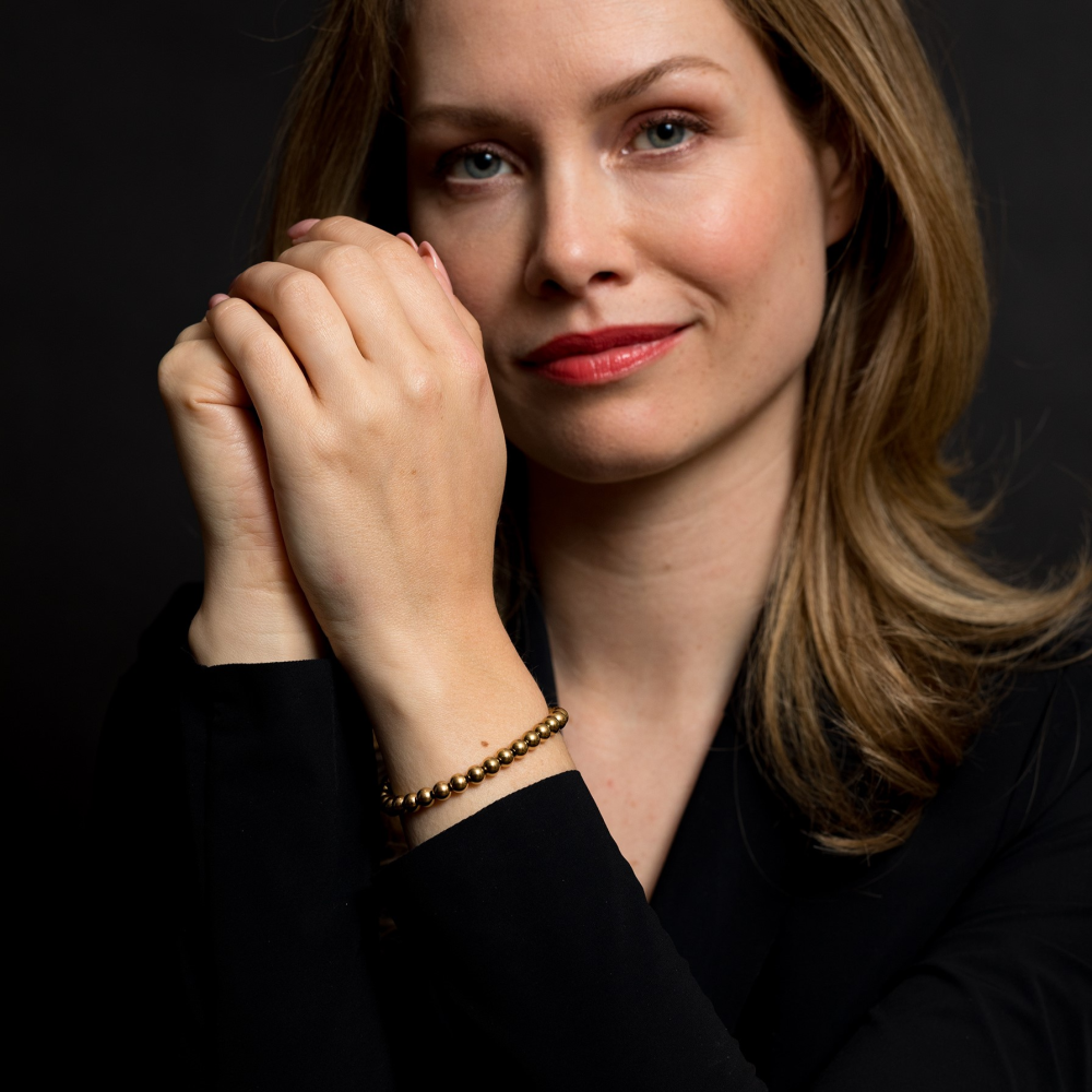 Een vrouw met lichtbruin haar en rode lippenstift, draagt een zwart topje en poseert met de handen bij haar gezicht geklemd. Ze showt de Rebel & Rose Yellow Gold Only 6mm Kralen Dames Armband tegen een donkere achtergrond.