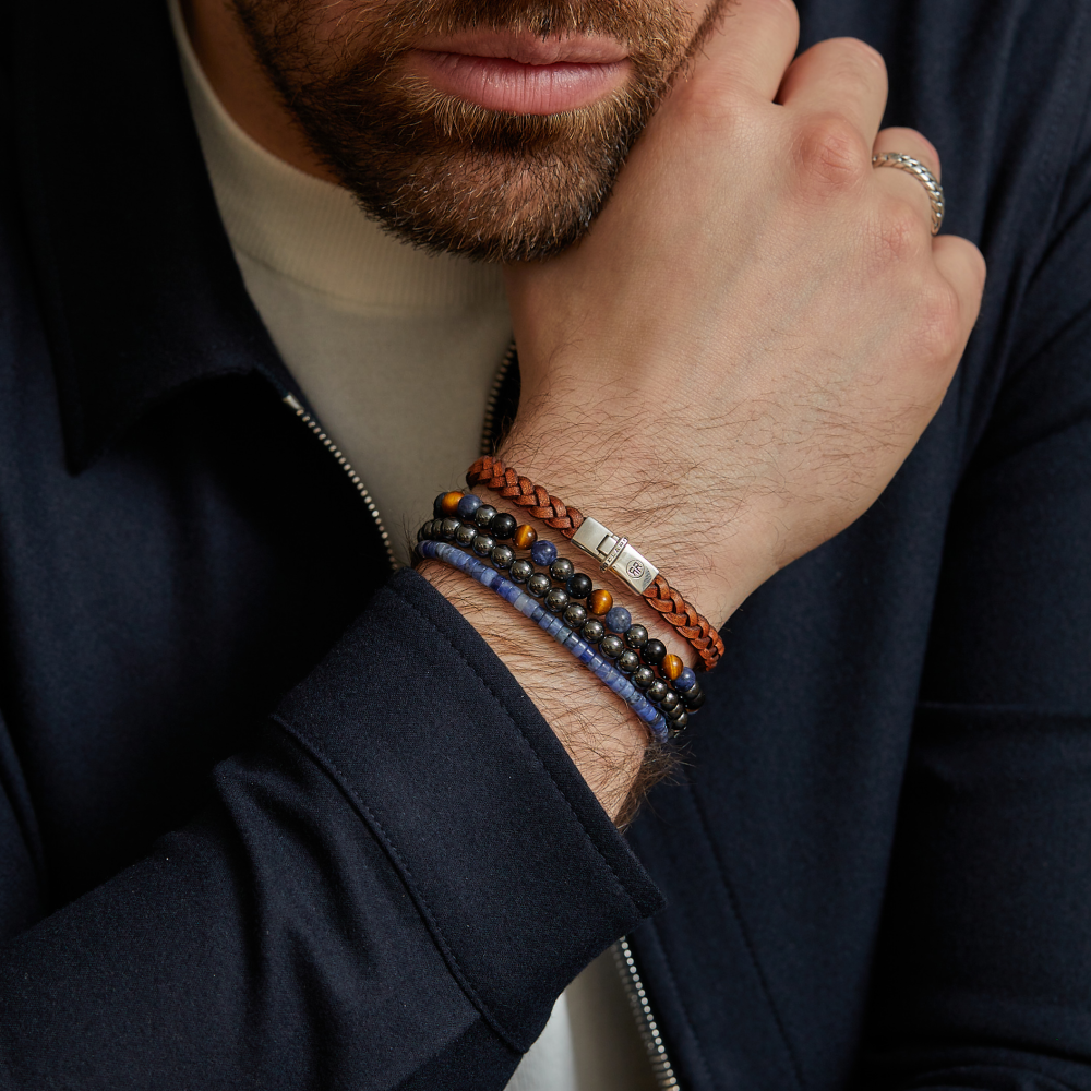 Een man met een getrimde baard draagt een donker jack en een crèmekleurig overhemd en laat zijn kin op zijn hand rusten. Hij draagt meerdere armbanden, waaronder de Rebel & Rose Grey Silver Shine DV 6mm heren armband, plus een zilveren ring om zijn vinger.