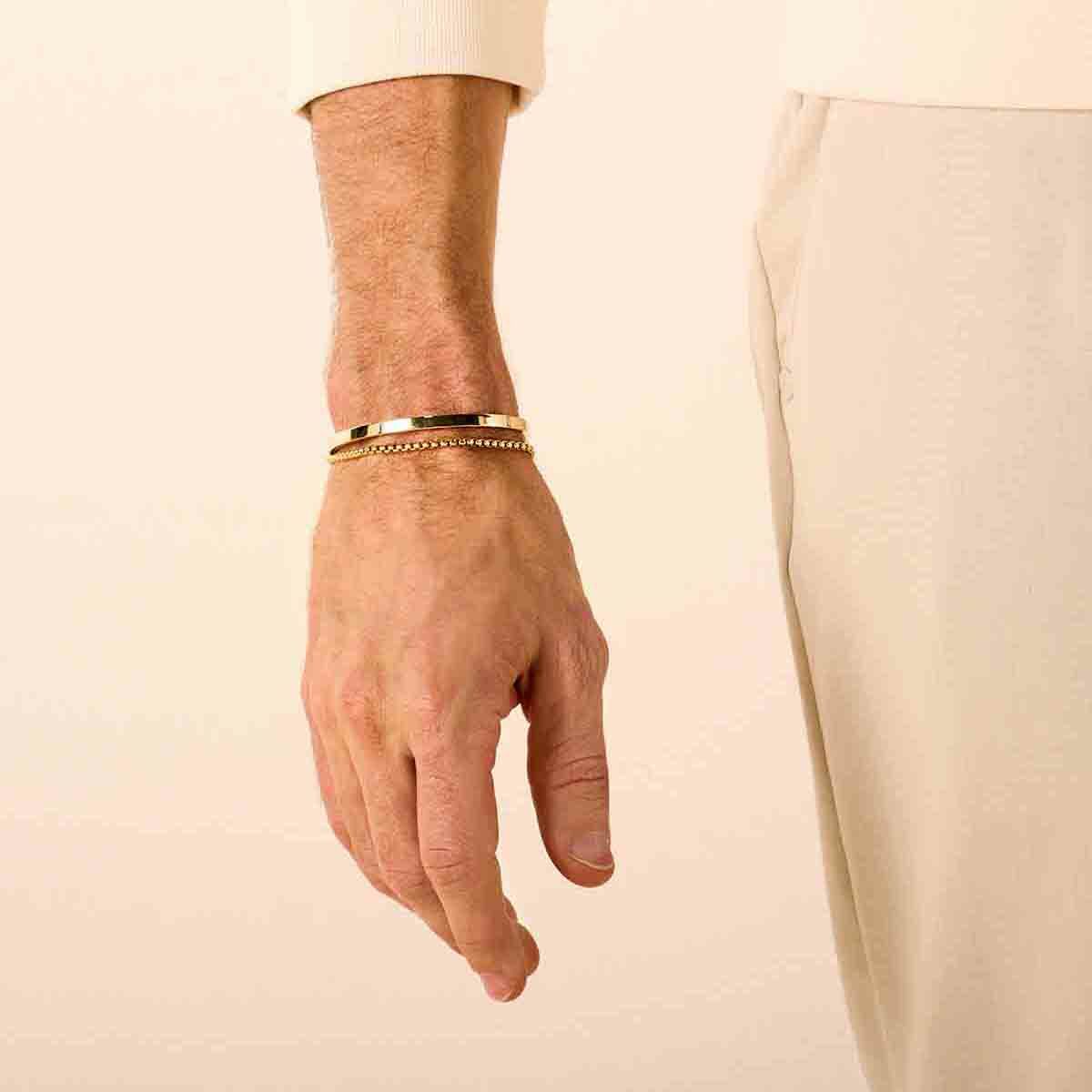 Een close-up van een linkerhand en -pols met twee gouden armbanden, waaronder de Van der Kooij Collectie bangle scharnier massief vlakke buis 4,0 mm Zilgold (geelgoud met zilverkern), gecombineerd met een crèmekleurig overhemd en beige broek tegen een effen achtergrond.