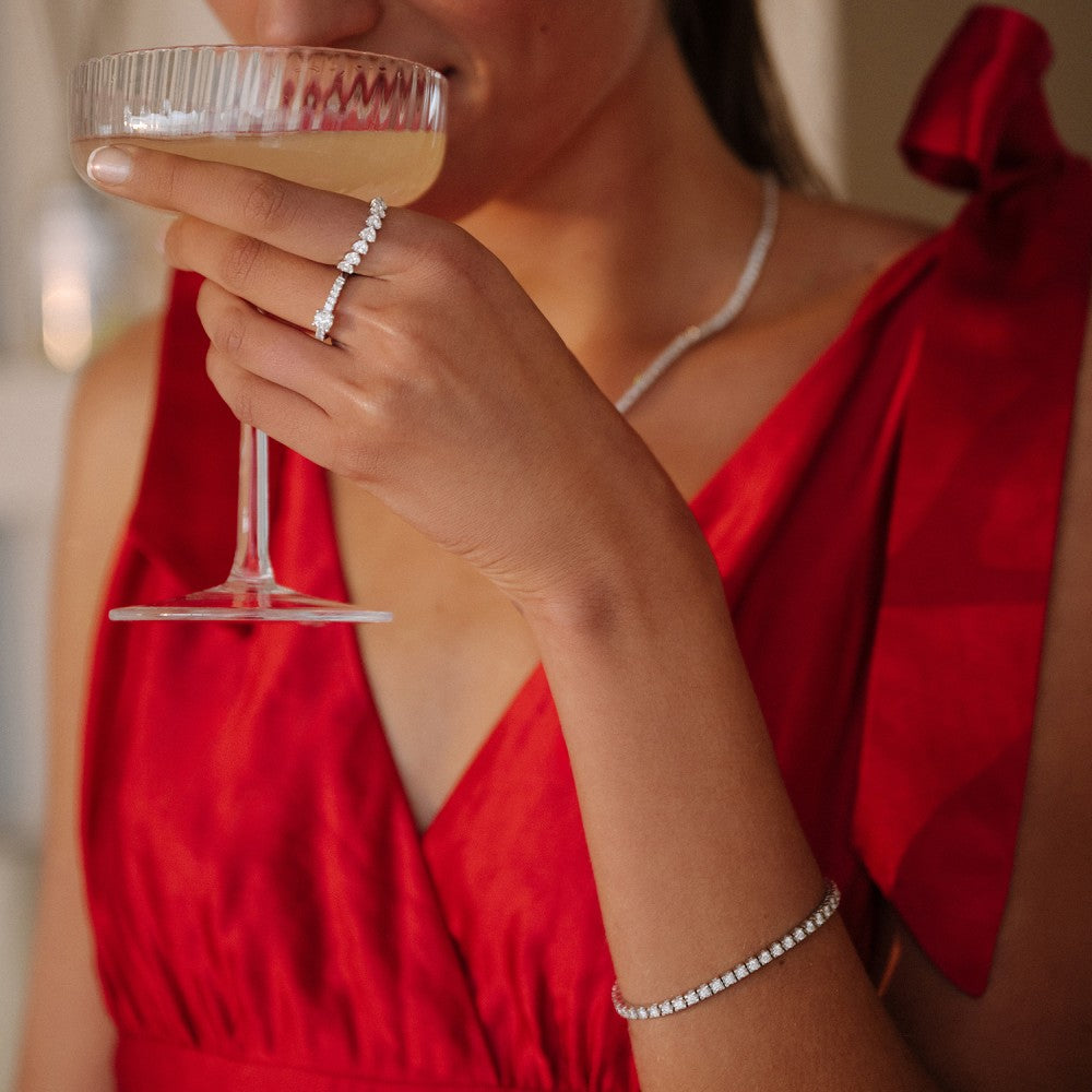 Een vrouw in een rode jurk houdt een glas champagne vast en pronkt met een diamanten ring, ketting en de armband Chloe 3.00 crt. geelgoud van Aurore Jewelry. Haar sieraden van 18 karaat gerecycled goud schitteren terwijl haar gezicht gedeeltelijk verborgen blijft.