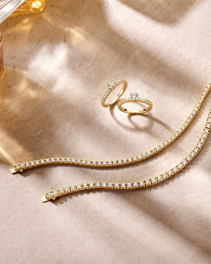 De Geelgouden Tennisarmband met 3.00 karaat diamanten in chatonzetting van Excellent Jewelry is elegant uitgestald op een beige, getextureerd oppervlak waarop zacht licht subtiele schaduwen werpt.