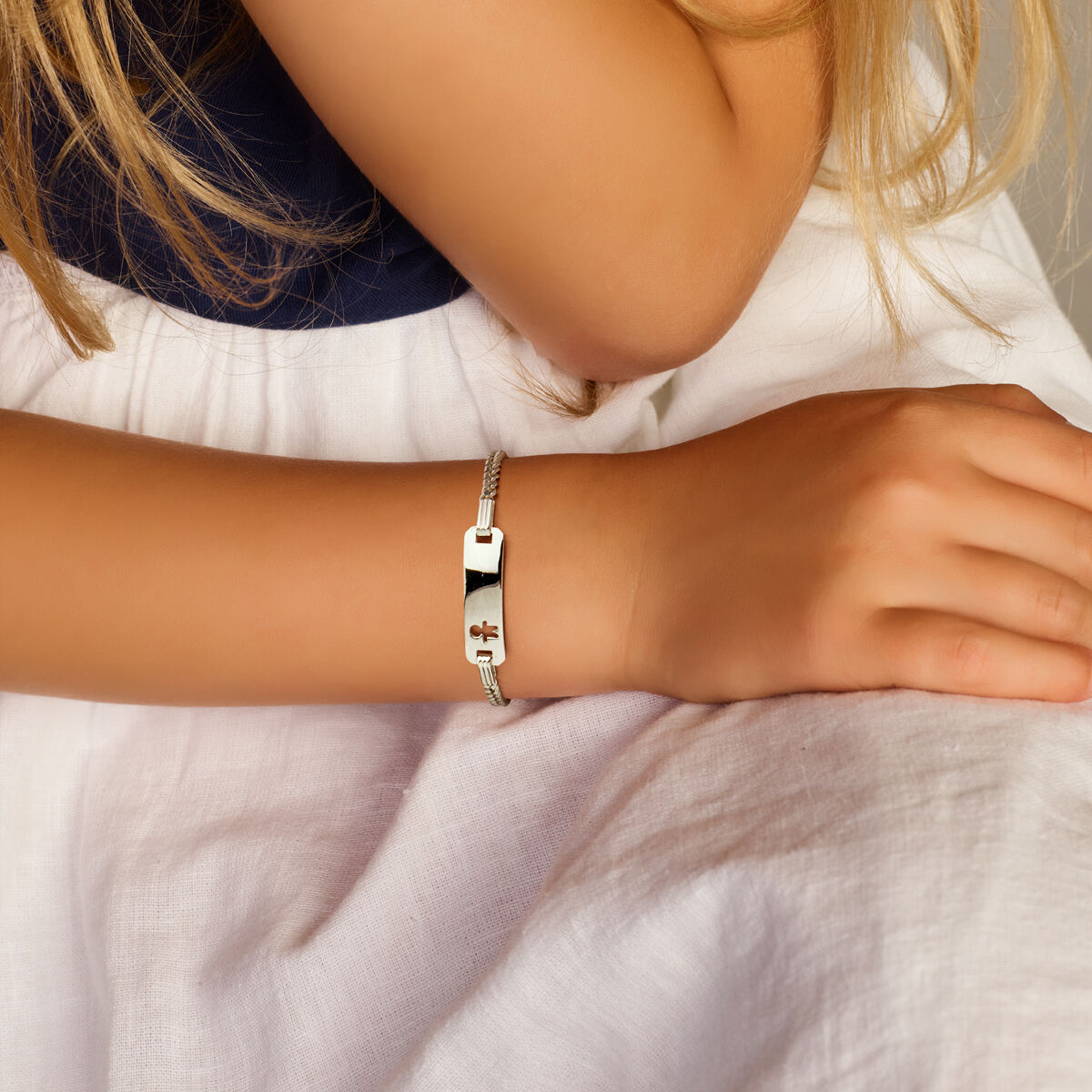 Een close-up toont de hand van een persoon op zijn schoot, die een Van der Kooij Collectie graveerarmband jongen gourmette plaat 5,5 mm zilver gerhodineerd met een kruisontwerp draagt. Gecombineerd met een blauwe top en witte rok, valt de armband op in dit elegante ensemble.