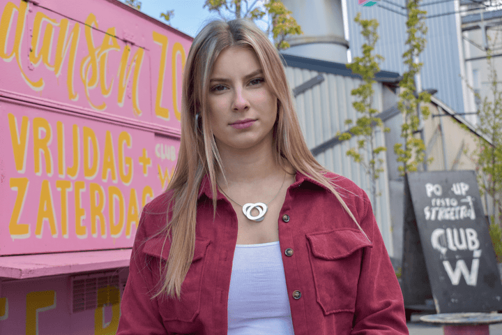 Een zelfverzekerde, stijlvolle vrouw met lang blond haar in een rood shirt staat bij een roze bord met gele tekst. Achter haar complementeren industriële elementen en een krijtbord haar unieke accessoire: de Collier Ovalen op elkaar Rood C70R van CLIC Creations—handgemaakt in Nederland.