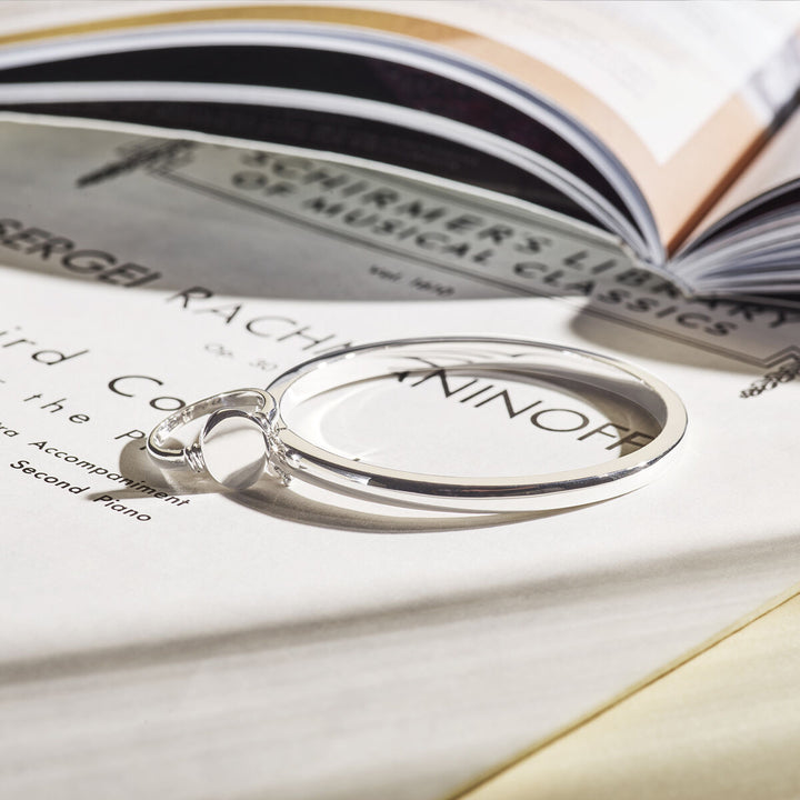 De bangle scharnier 4 mm bolle buis zilver gerhodineerd van Van der Kooij Collectie ligt op open bladmuziek en een boek, terwijl zacht licht subtiele schaduwen werpt.