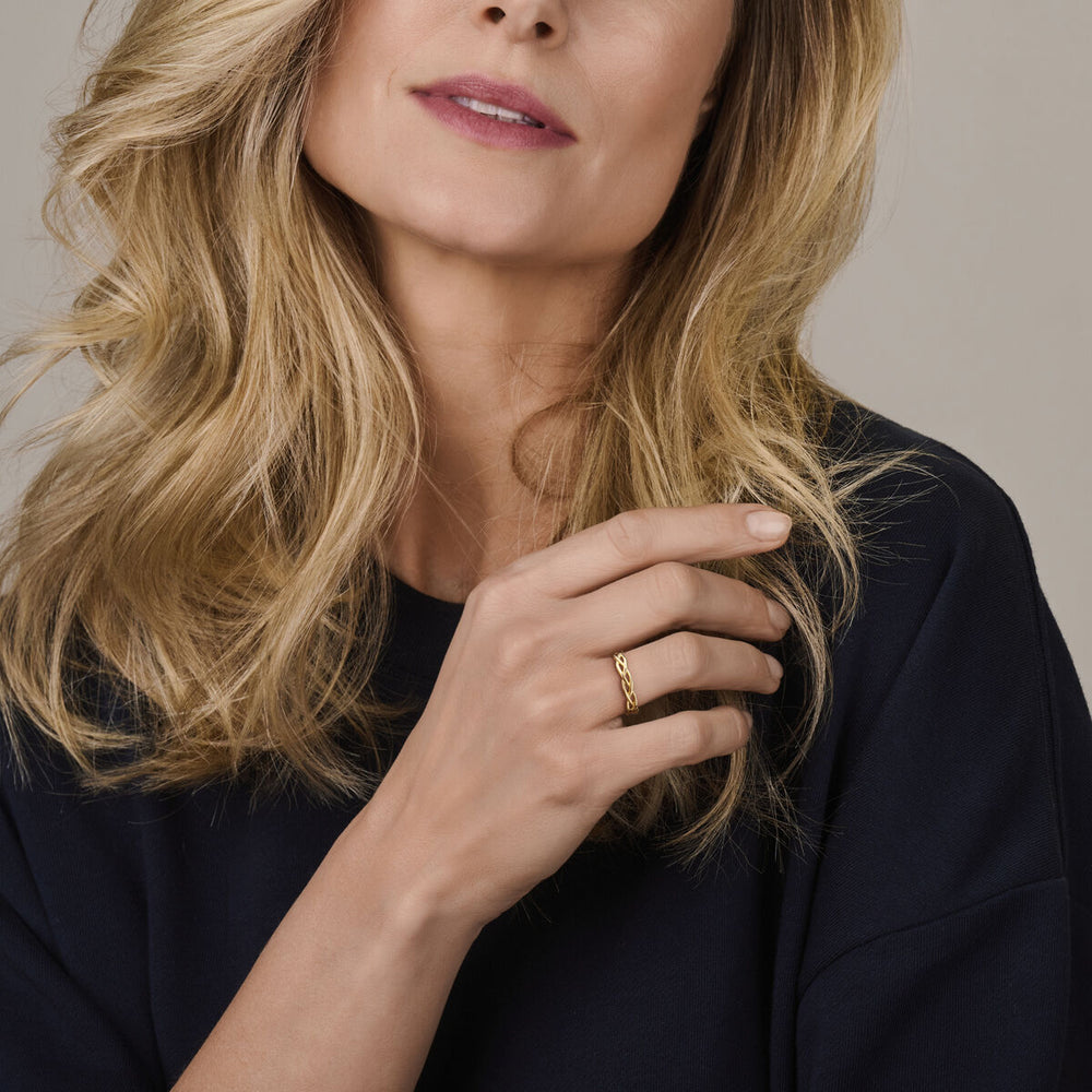 Een vrouw met golvend blond haar in een donker topje poseert met haar hand voor haar gezicht. Ze showt de Van der Kooij Collectie ring gevlochten 14K geelgoud - tijdloze elegantie in een gevlochten gouden ontwerp.