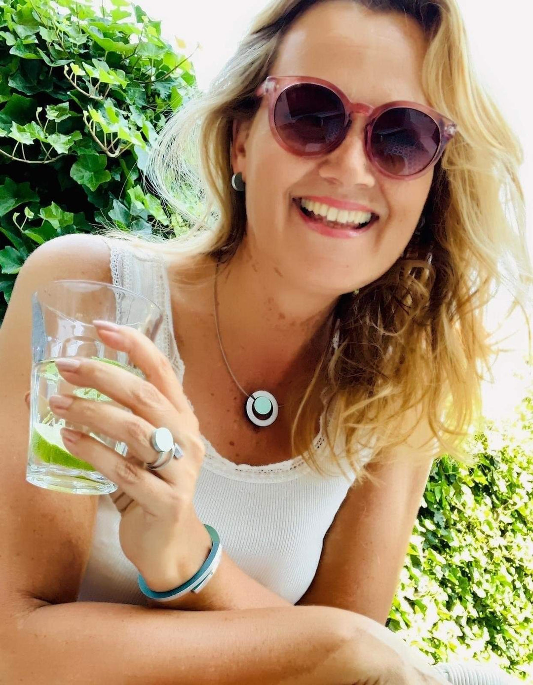 Een vrouw met lang blond haar lacht, houdt een met limoen versierd drankje vast en draagt een stijlvolle zonnebril, een wit topje en de elegante CLIC Creations Ring Twee ronde vormen Grijs R5AG. Groen gebladerte siert de achtergrond terwijl zonlicht de scène opfleurt.